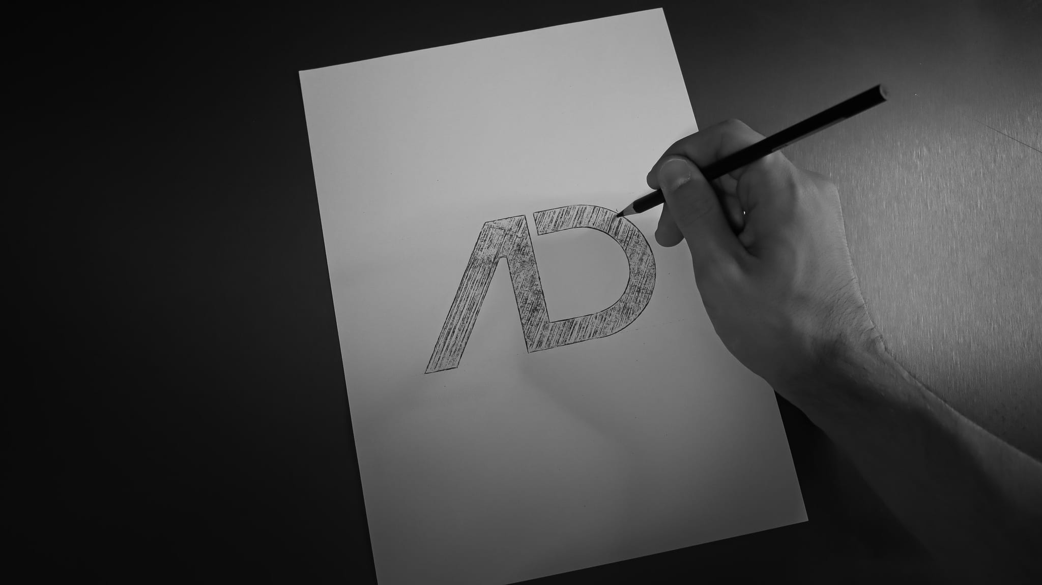 Una mano disegna a matita il monogramma AD su un foglio bianco, riempiendo le due lettere di tratteggio: il foglio sta su una scrivania scura.
