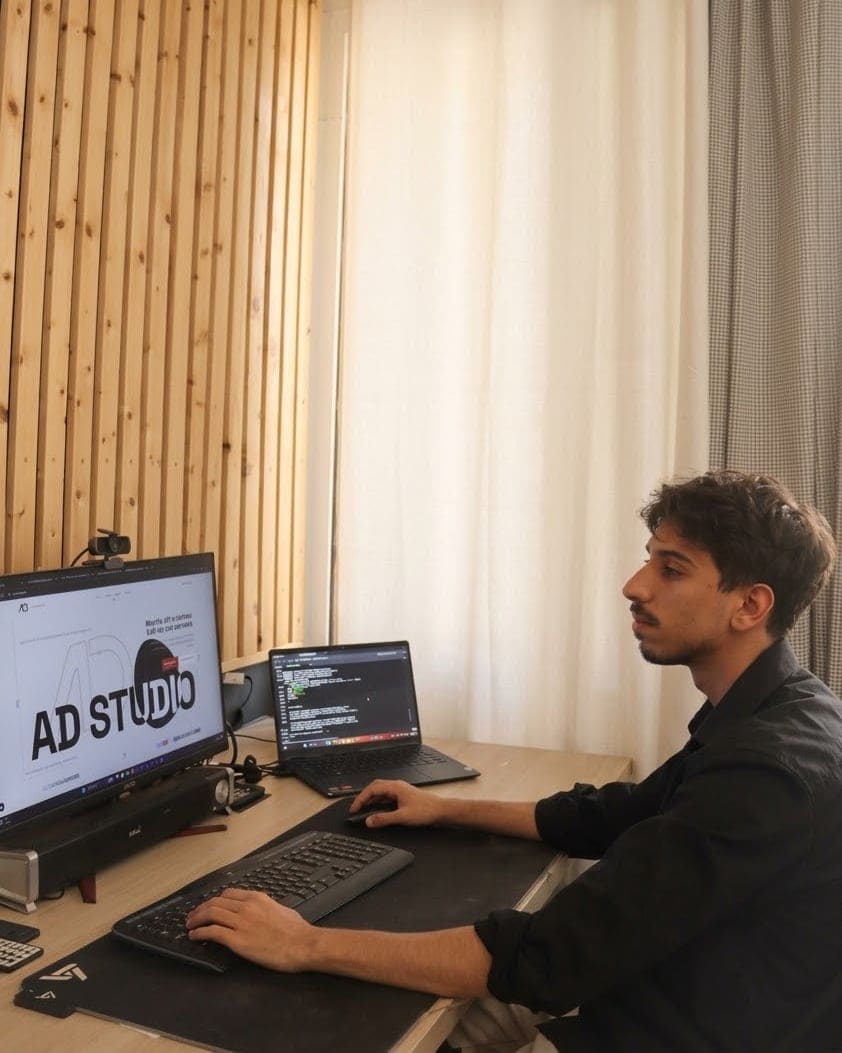 Dennis alla scrivania, di profilo, una mano sulla tastiera e una sul mouse. Sullo schermo grande c’è la pagina di apertura di questo sito, sul portatile accanto delle righe di codice.